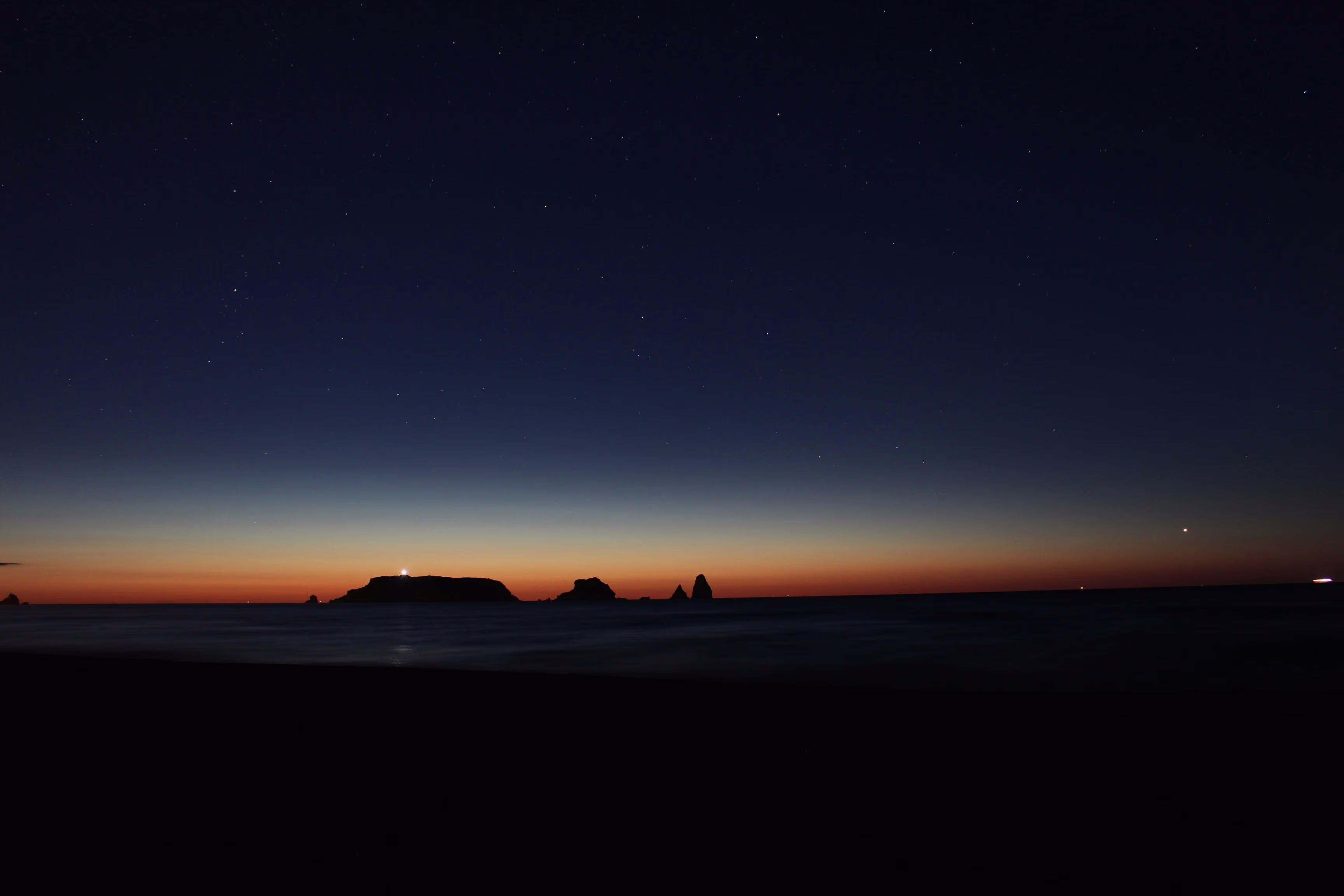 Horitzó marí a l'hora blava amb un cel estrellat, amb un far brillant a l'illa fosca a la distància.