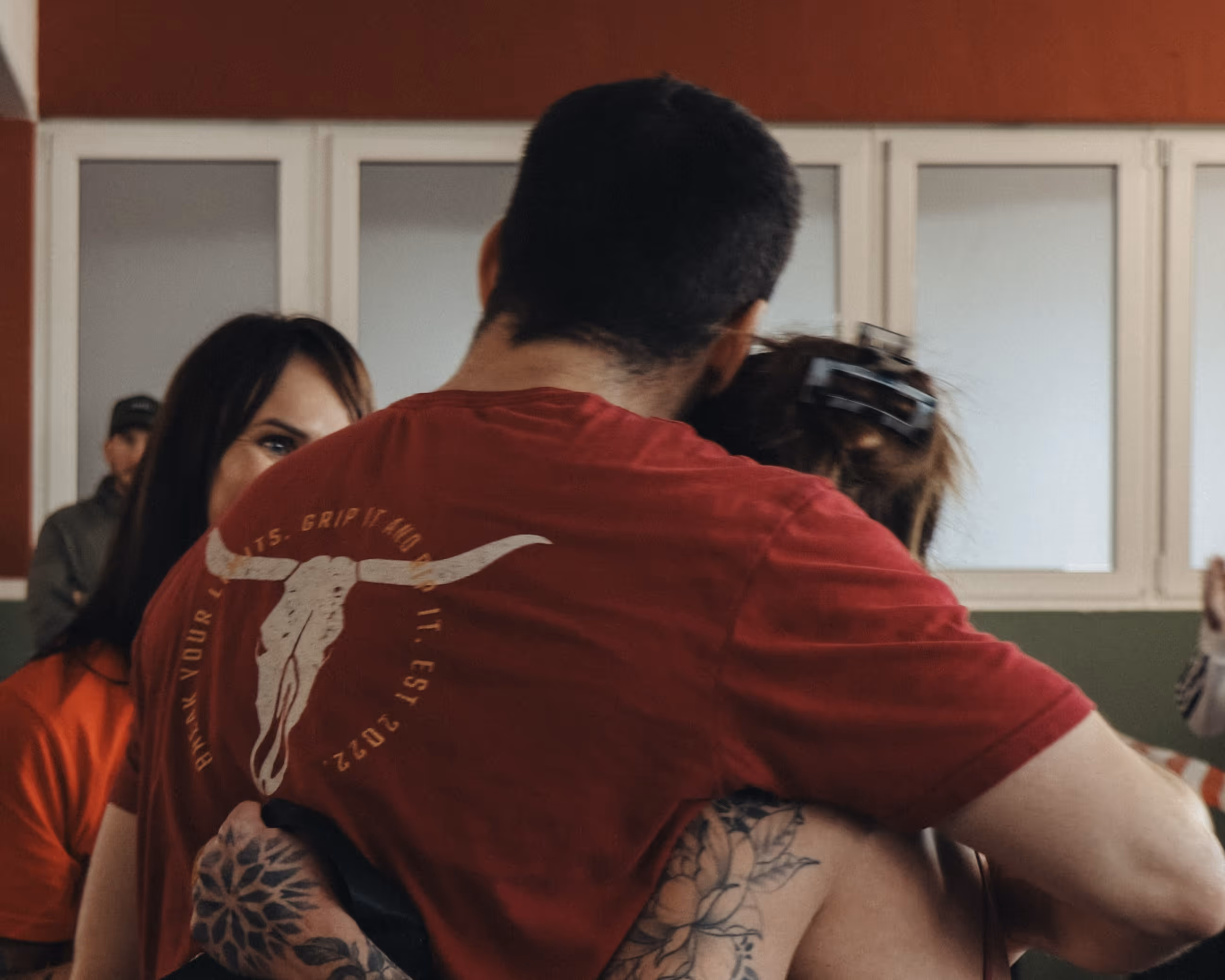 Person in rotem T-Shirt mit Stierschädel-Motiv umarmt eine andere Person mit Tattoo am Arm.