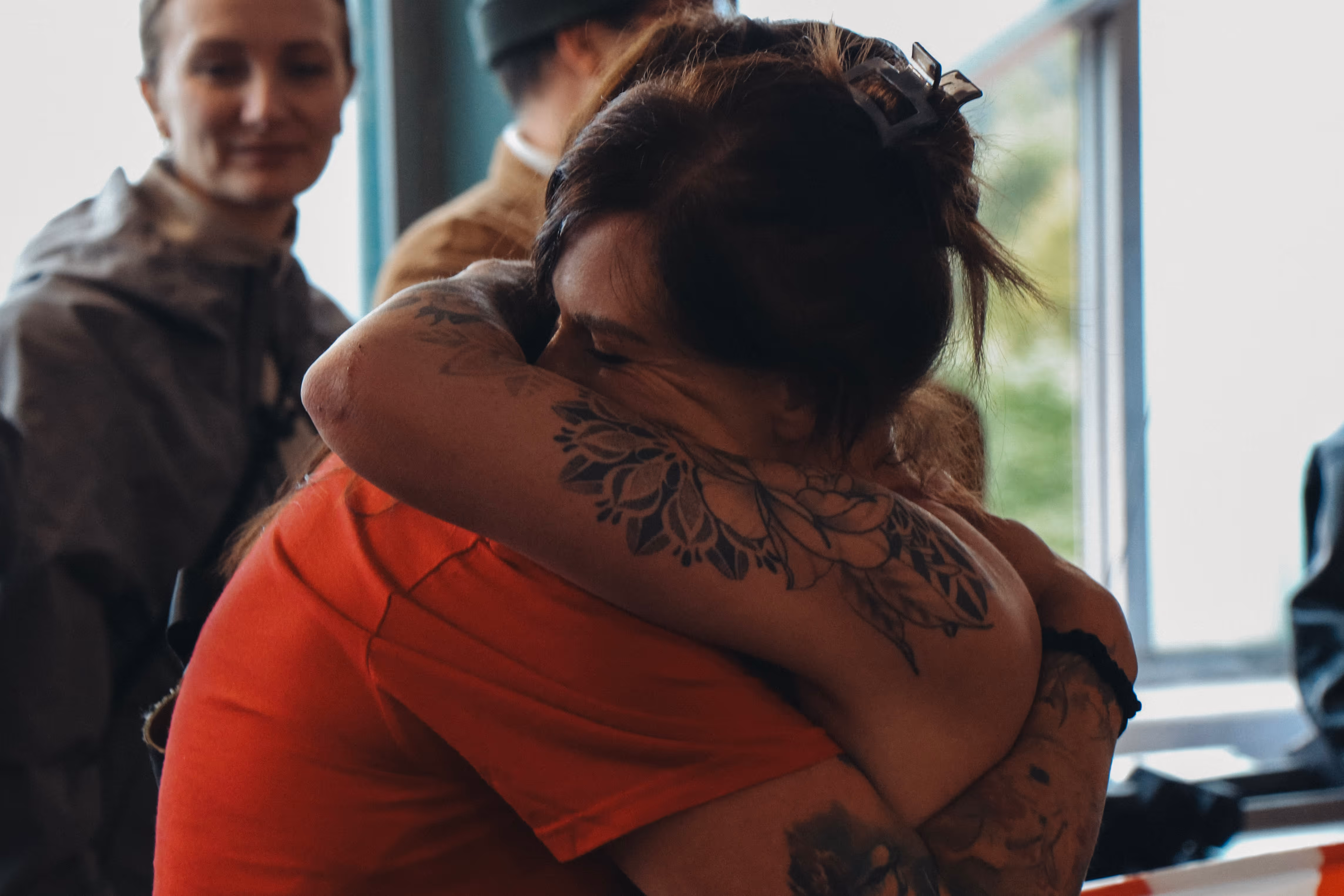 Zwei Personen umarmen sich herzlich, eine davon mit auffälligen Tätowierungen auf dem Arm und im Haar eine Haarspange.