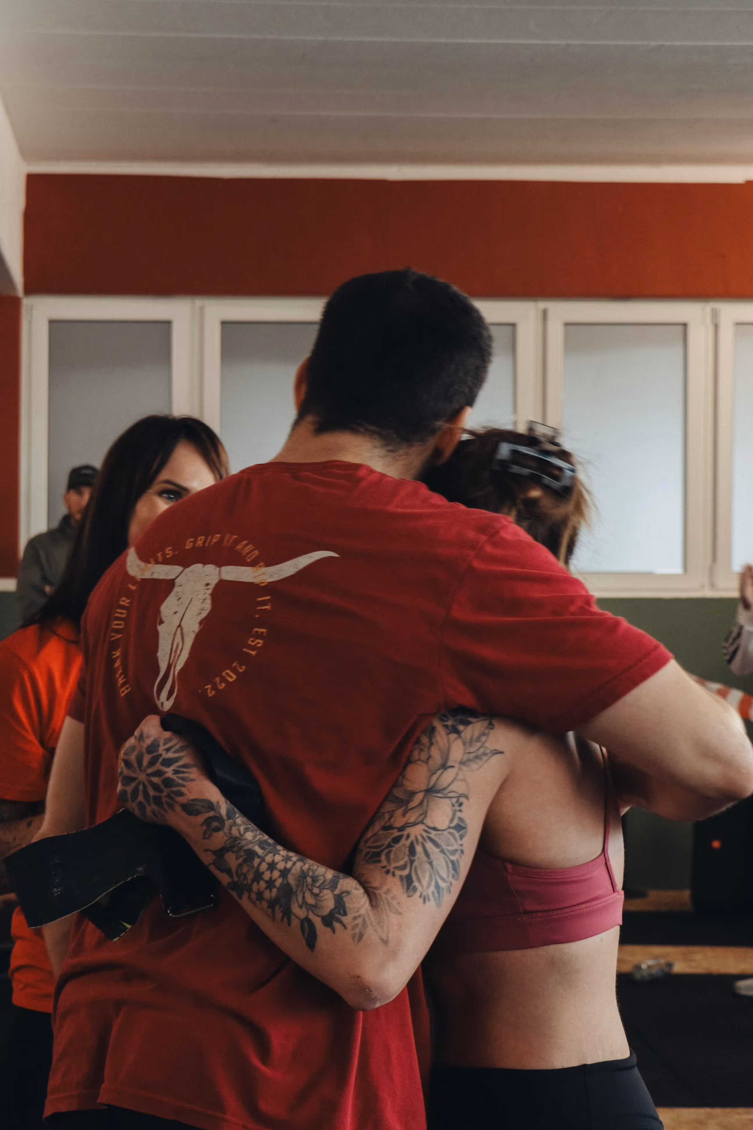 Zwei Menschen in Sportkleidung umarmen sich in einem Innenraum, eine Person hat ein florales Tattoo auf dem Arm.