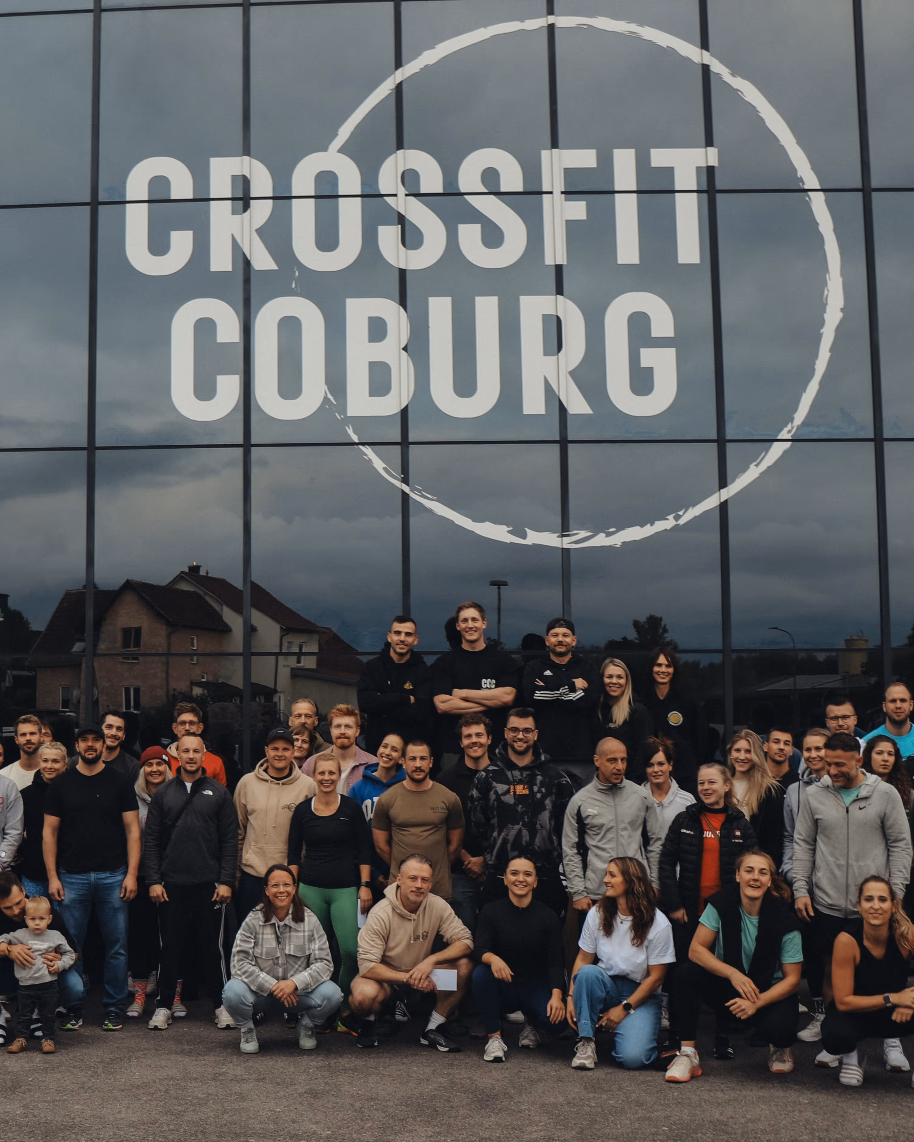 Gruppenfoto von Männern, Frauen und einem Kind vor einer Glasfassade mit der Aufschrift 'Crossfit Coburg'.