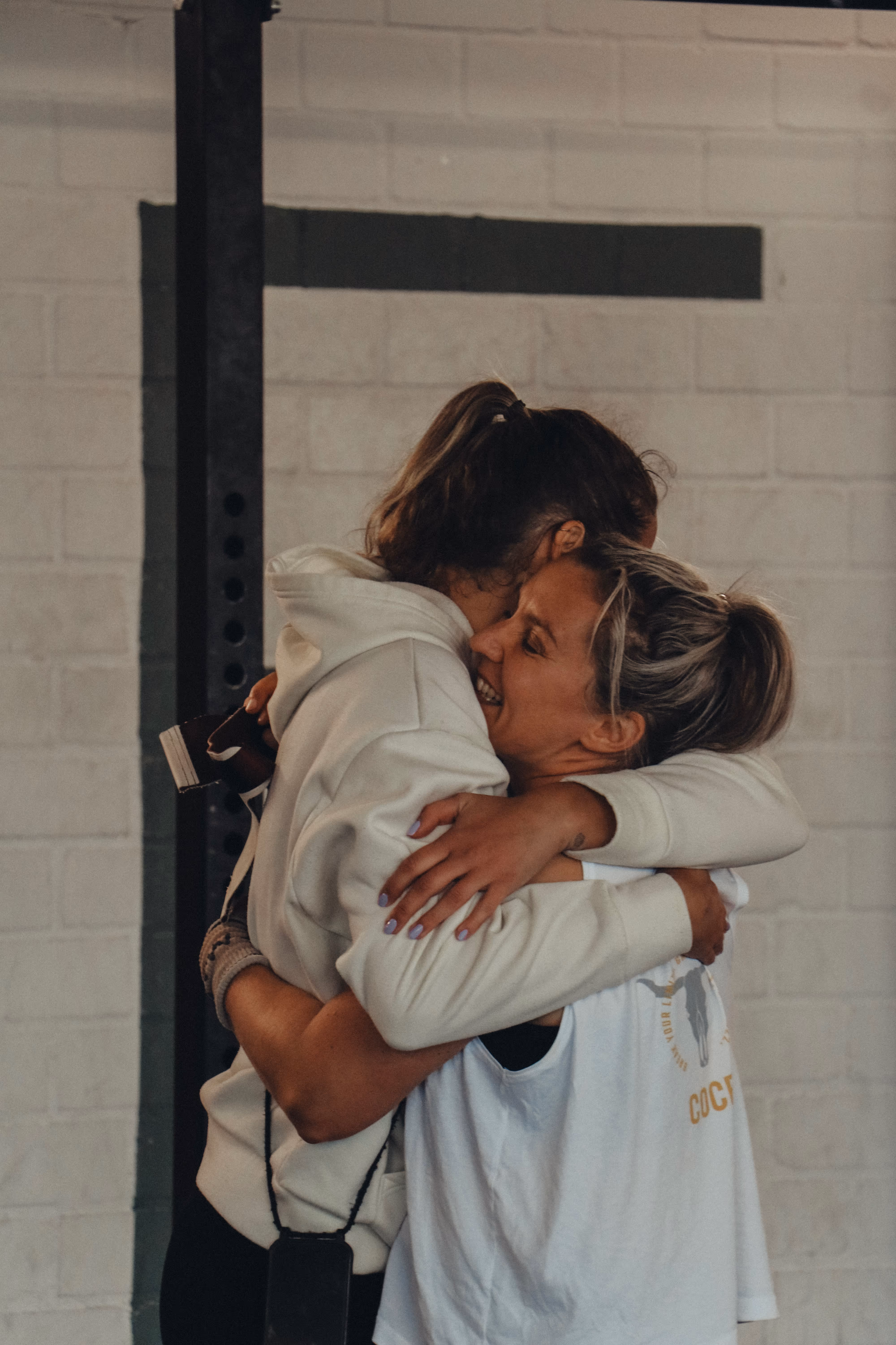 Zwei Frauen umarmen sich in einem Fitnessstudio vor einer weißen Backsteinwand.