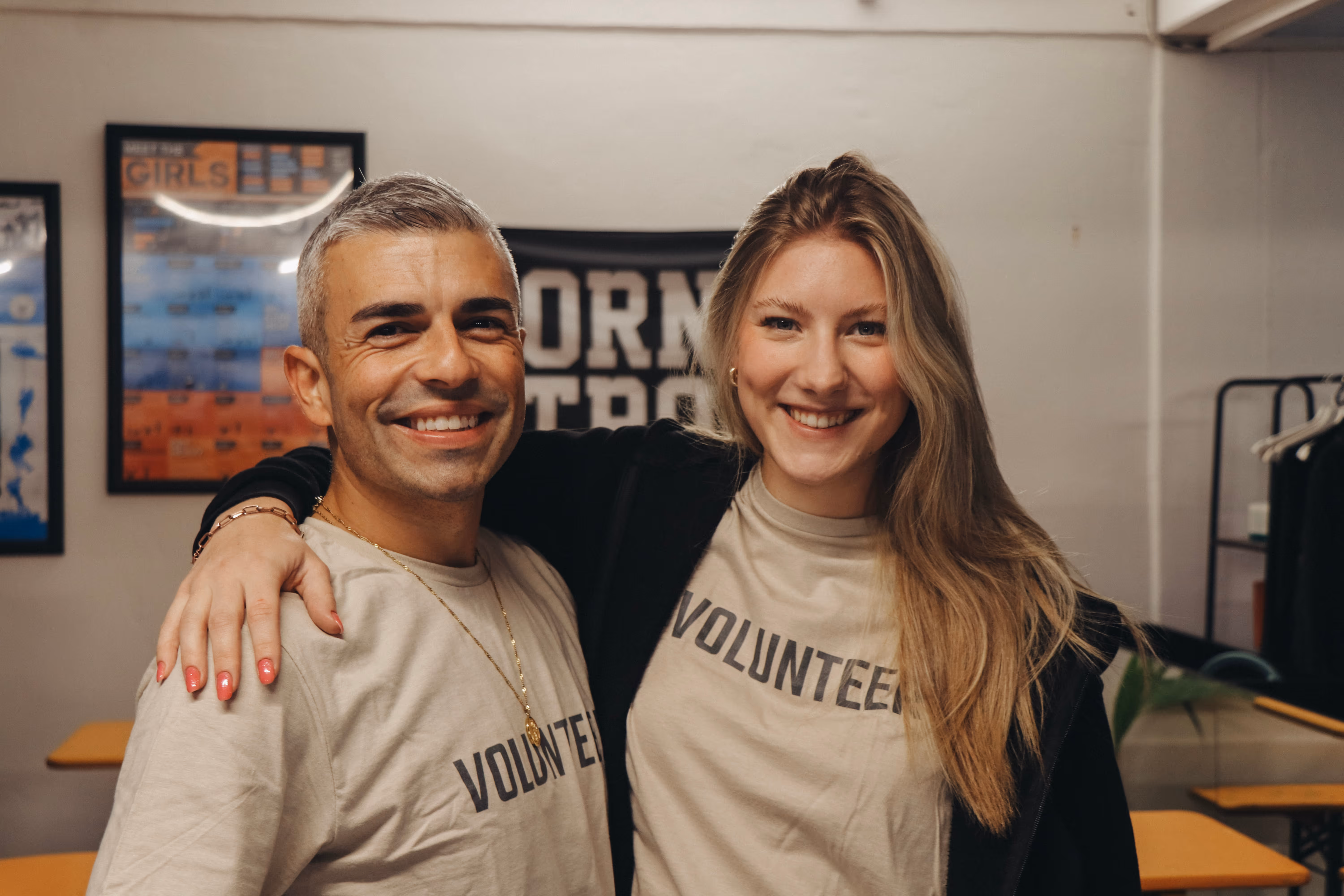 Zwei lächelnde Freiwillige mit beigen T-Shirts, auf denen "VOLUNTEER" steht, posieren eng zusammen.
