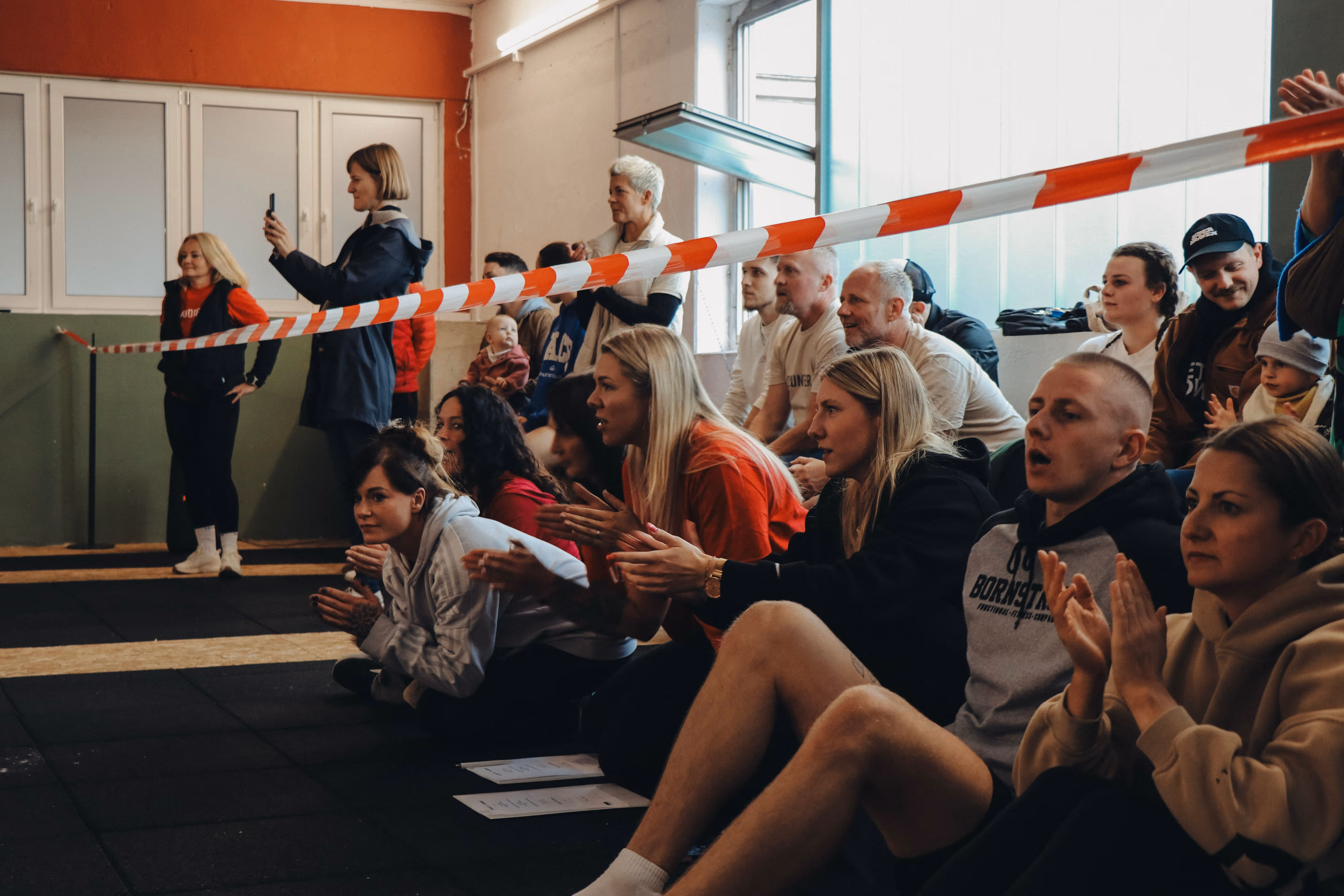 Zuschauer in einer Halle, einige sitzen und klatschen hinter einem rot-weißen Absperrband, eine Frau macht ein Foto mit dem Handy.