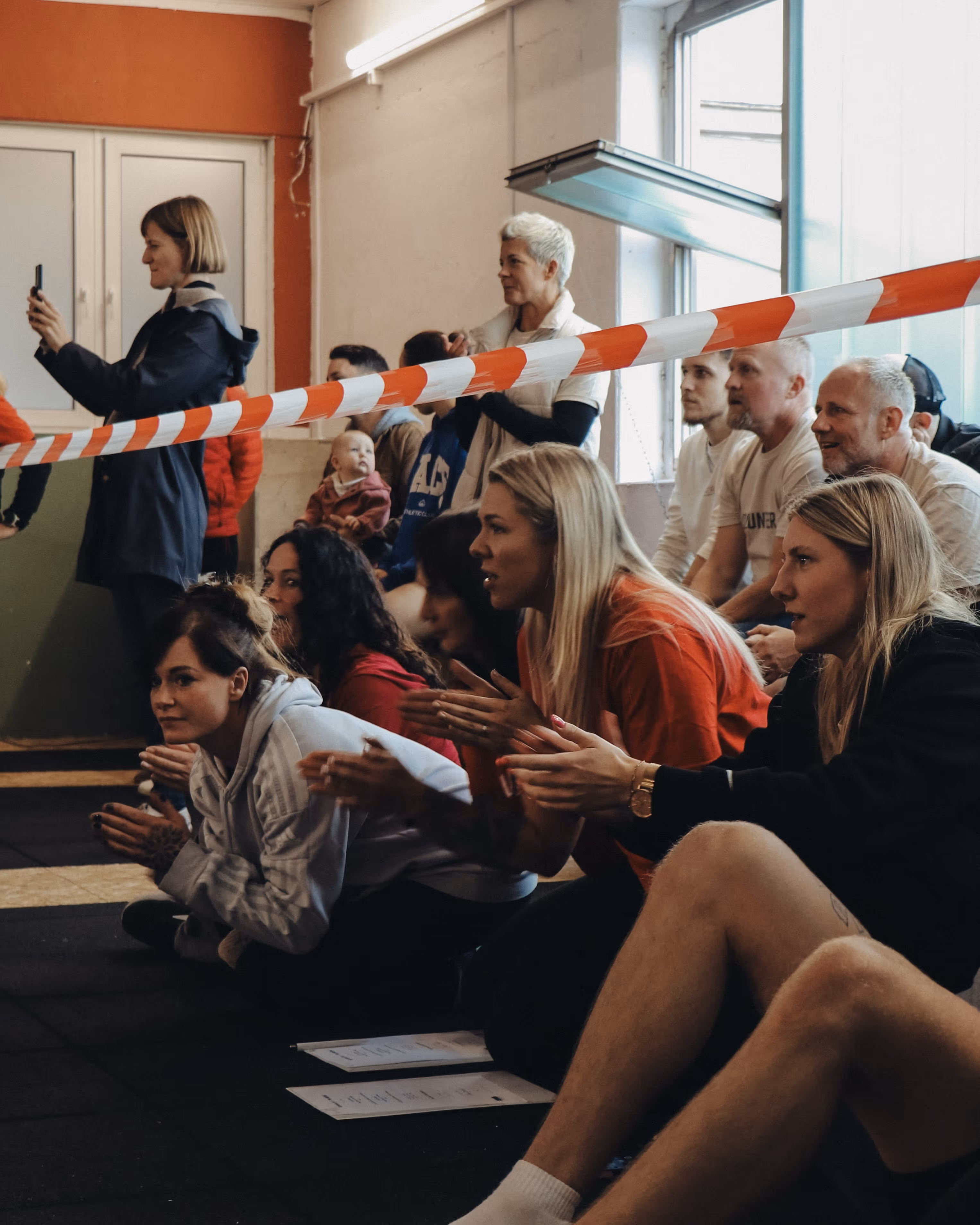 Zuschauer in einer Halle, einige sitzen und klatschen hinter einem rot-weißen Absperrband, eine Frau macht ein Foto mit dem Handy.