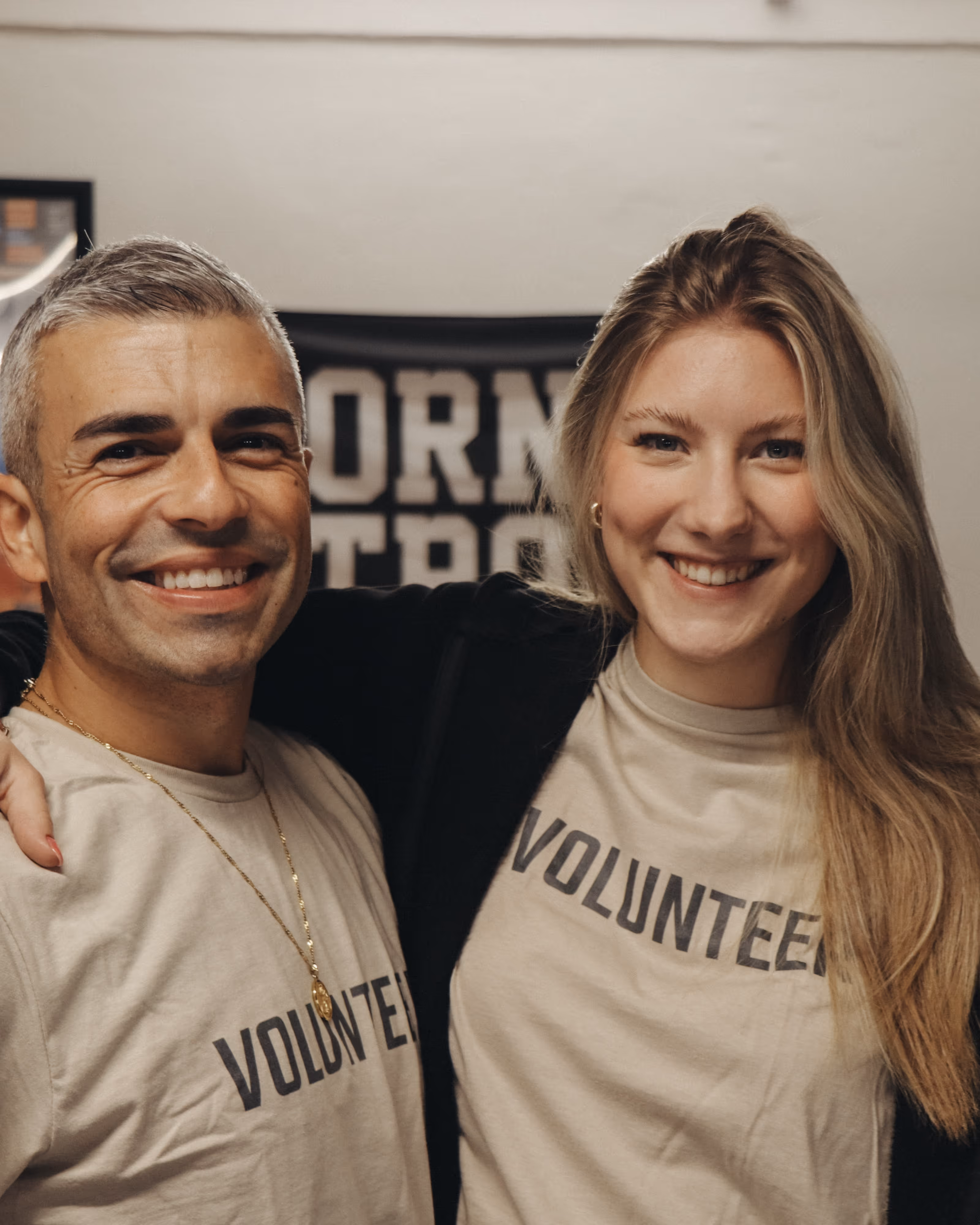 Zwei lächelnde Freiwillige mit beigen T-Shirts, auf denen "VOLUNTEER" steht, posieren eng zusammen.