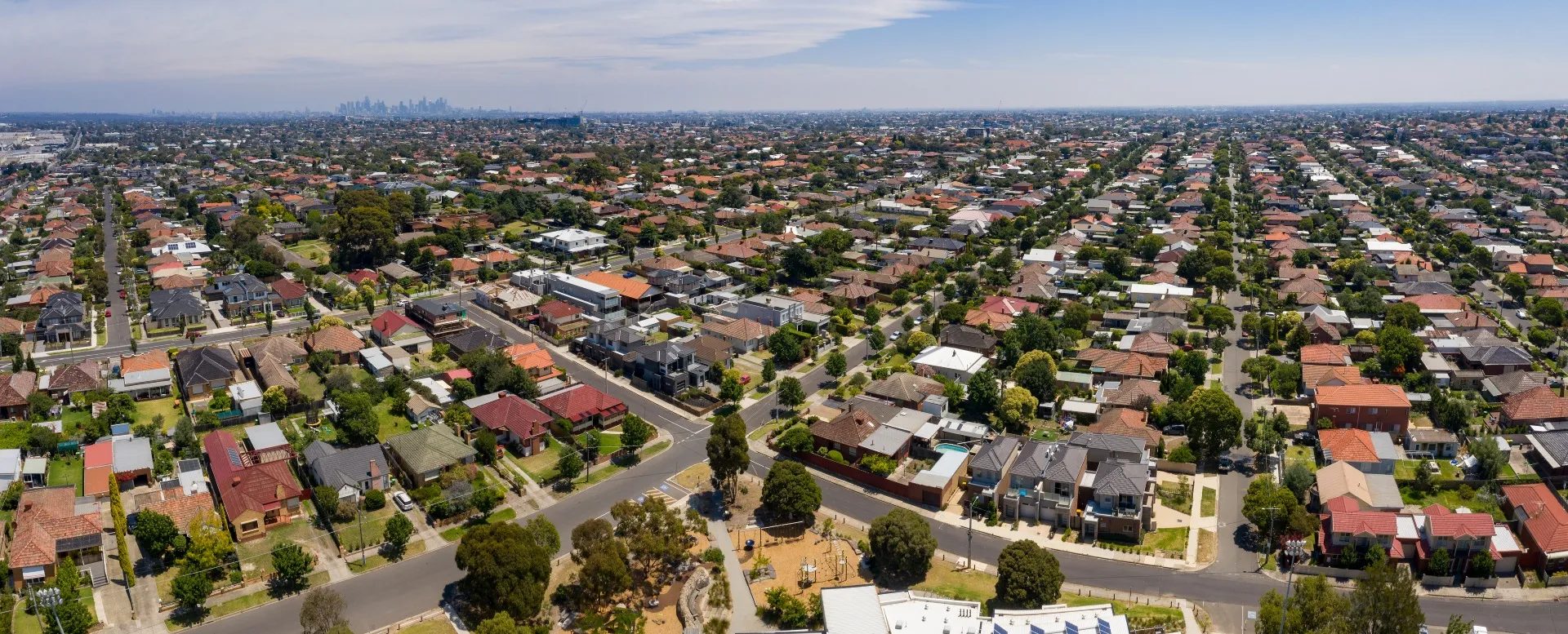 Aerial view of an australian suburban neighborhood with numerous houses, tree-lined streets, and a city skyline in the distance under a partly cloudy sky.