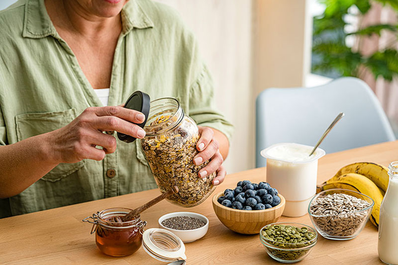 Photo of woman opening a glass jar of granola. blueberries, seeds and yogurt on the table.