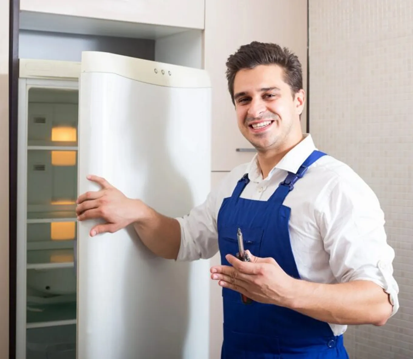 Smiling technician in blue overalls opening a white refrigerator door while holding a tool.