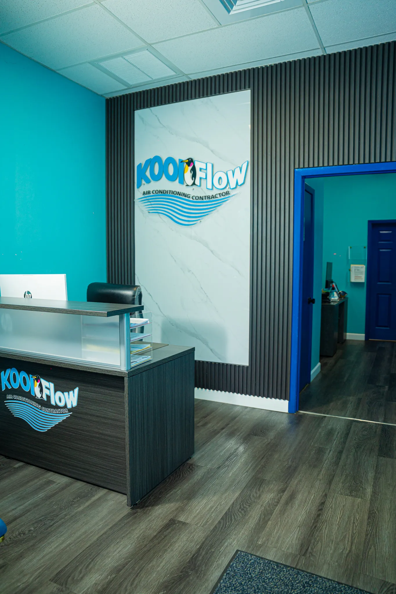Office reception area with a dark wood desk displaying KOOLFlow air conditioning contractor logo, turquoise walls, and a doorway leading to an interior room.