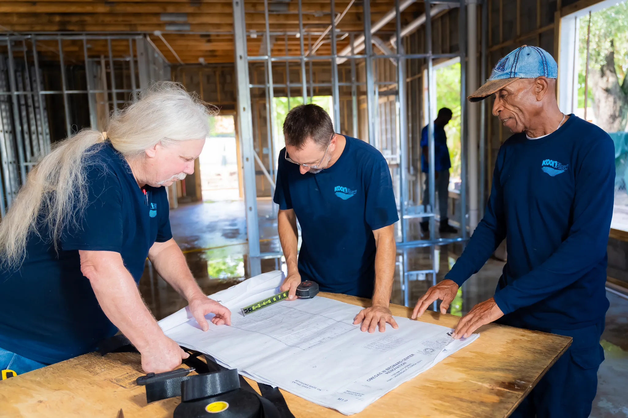 Three construction workers in blue shirts reviewing blueprints on a wooden table inside an unfinished building.