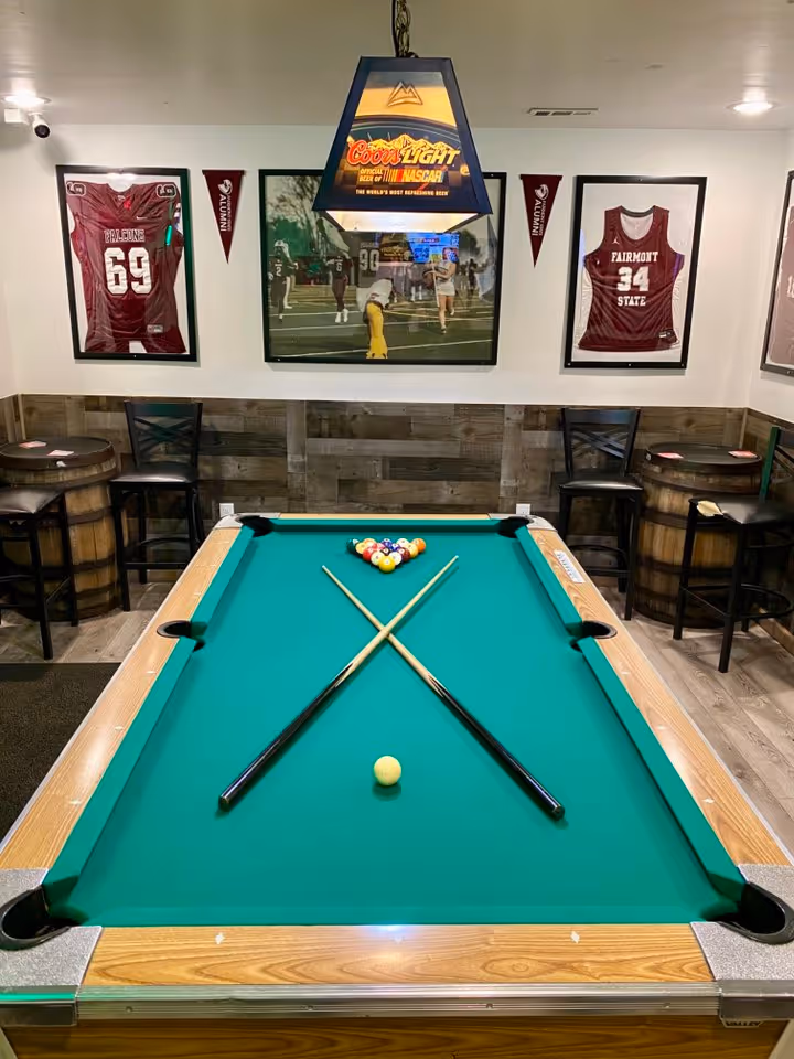 A dimly lit billiard room with a pool table at the center, crossed cues, and a racked set of balls.