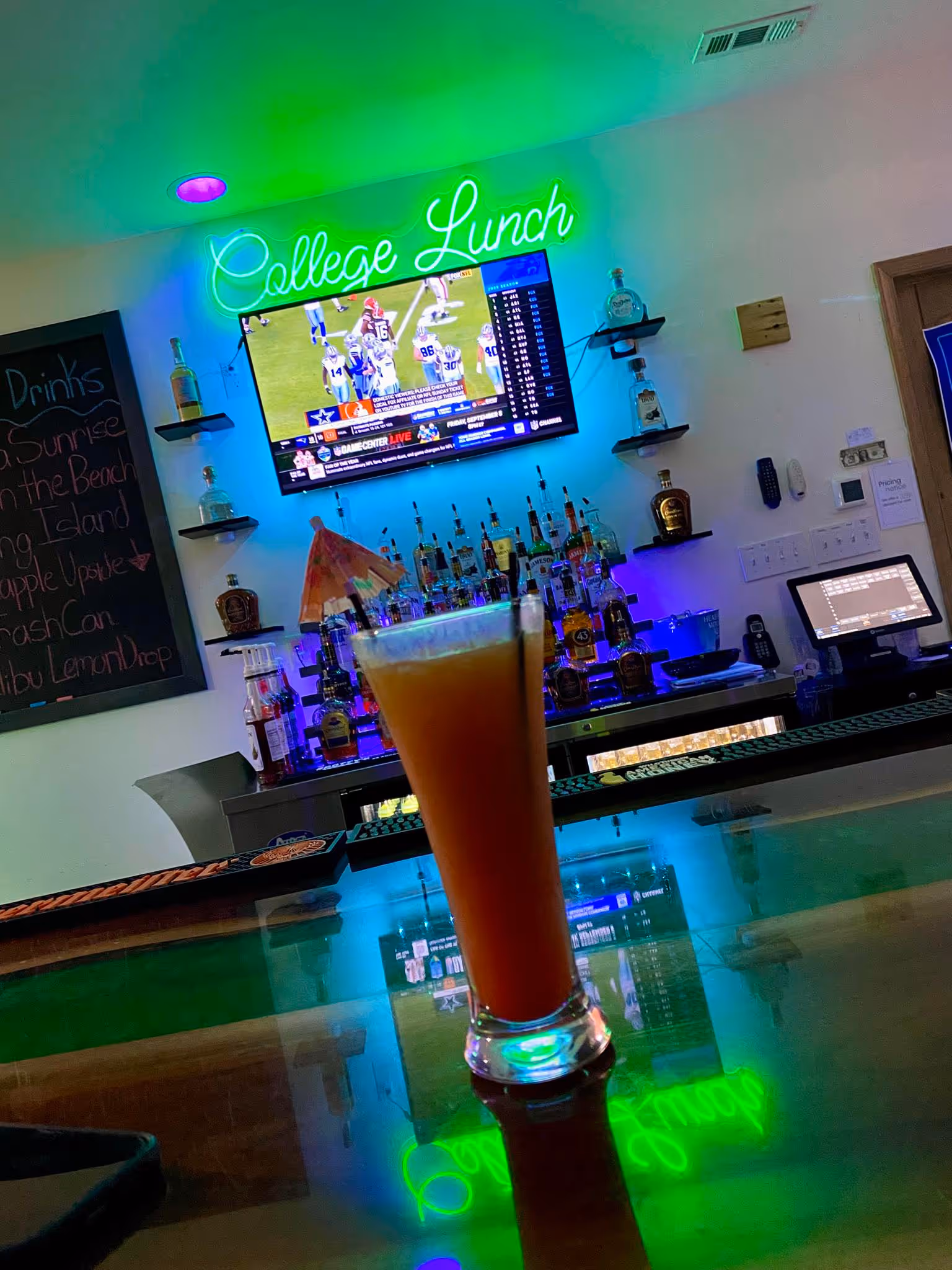 A tall orange cocktail on a bar counter, under a neon "College Lunch" sign.