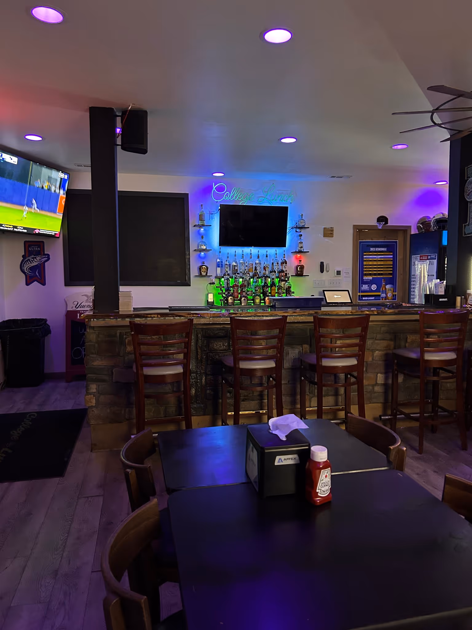 A cozy bar with wooden stools, colorful backlit shelves of liquor, neon lights, and a TV.