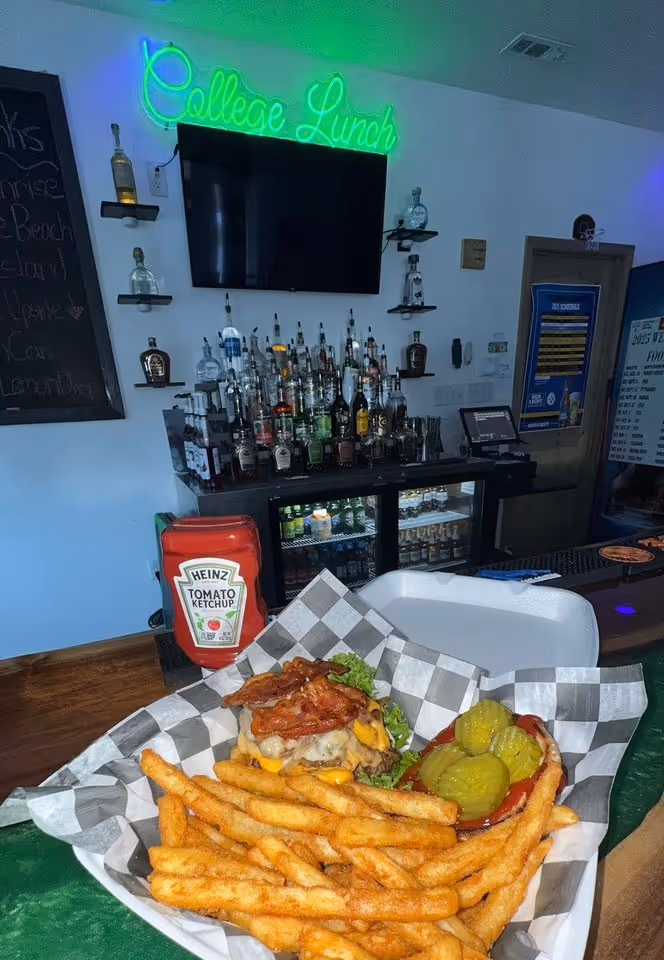 A juicy burger with bacon, cheese, lettuce, and pickles, served with crispy fries in a checkered basket, sits on a bar counter. In the background, a neon "College Lunch" sign glows above a stocked bar with various liquor bottles. The atmosphere is casual and inviting.