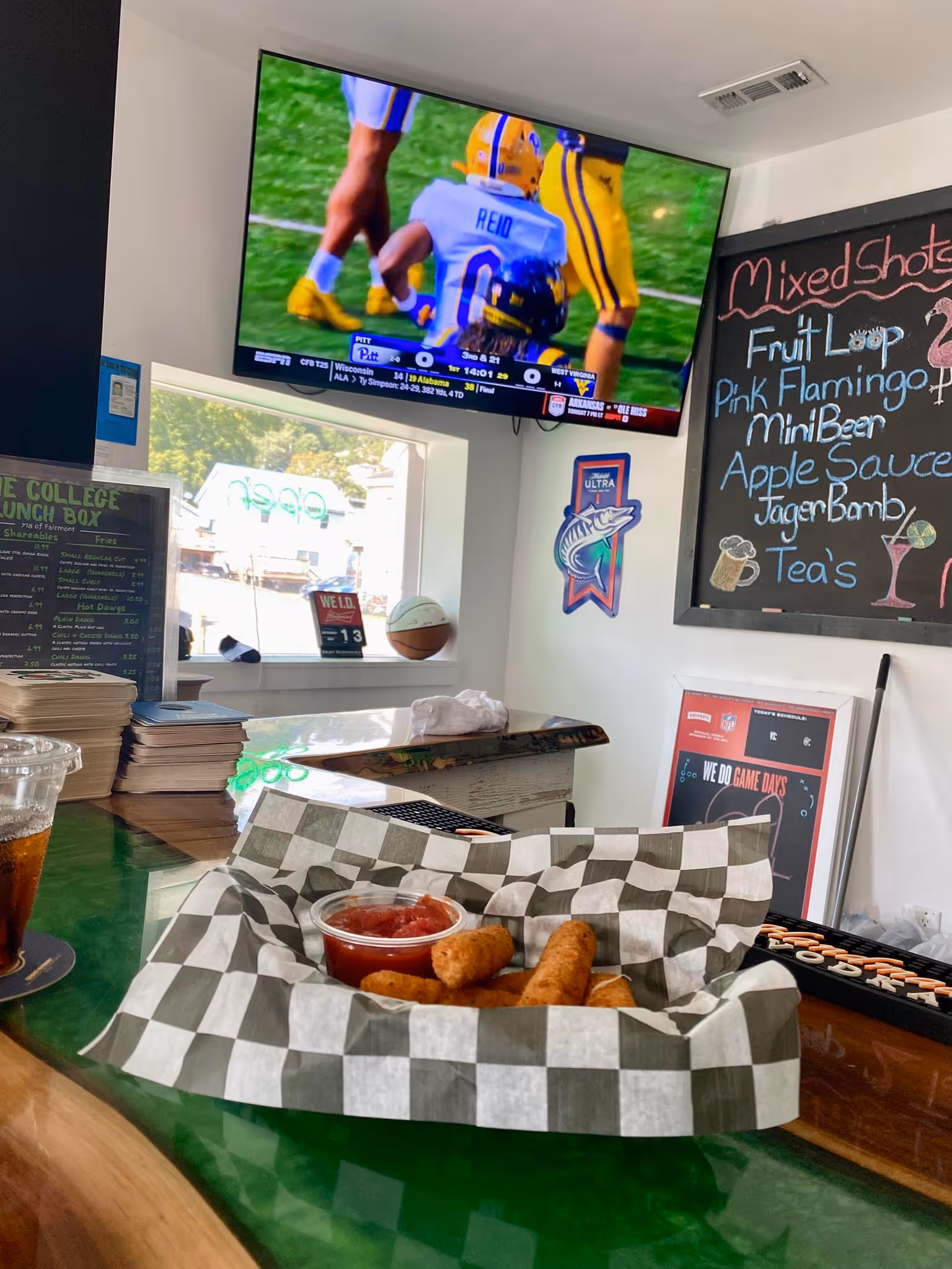 A bar counter with a checkered basket of mozzarella sticks and sauce in the foreground. In the background, there is a TV showing a football game; a chalkboard lists drink specials. 