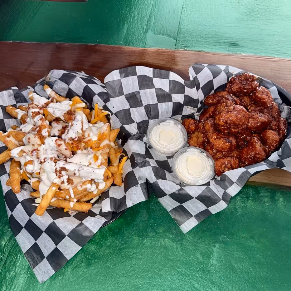 Two checkered baskets on a green table with loaded fries topped with cheese and a creamy sauce on the left, and saucy chicken wings with dipping cups on the right.