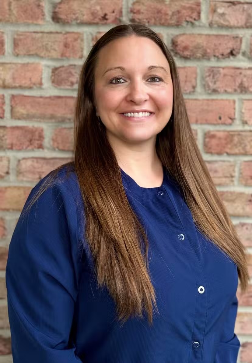 Smiling woman with long brown hair wearing a blue buttoned shirt standing in front of a brick wall.