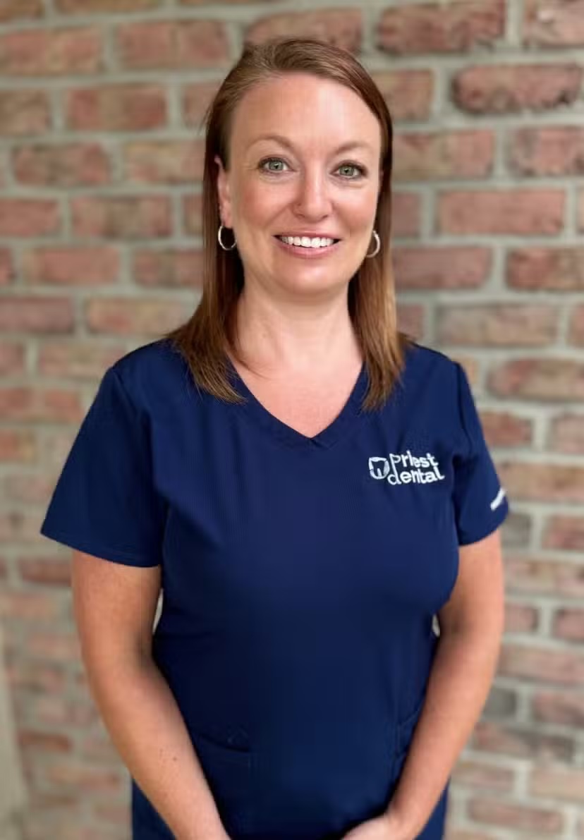 Smiling woman with straight brown hair wearing a navy blue Priest Dental scrub top standing in front of a brick wall.