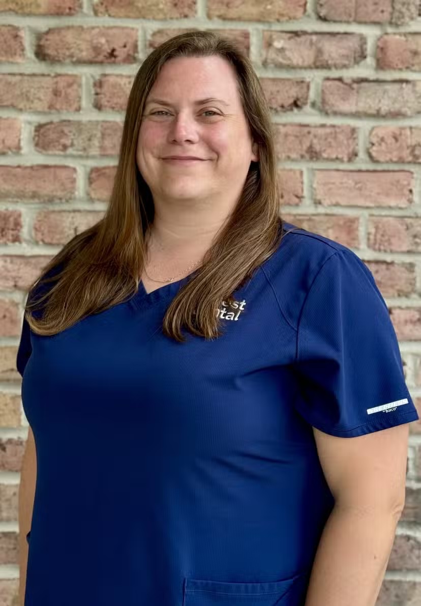 Smiling woman with long brown hair wearing a blue scrub top standing against a brick wall.