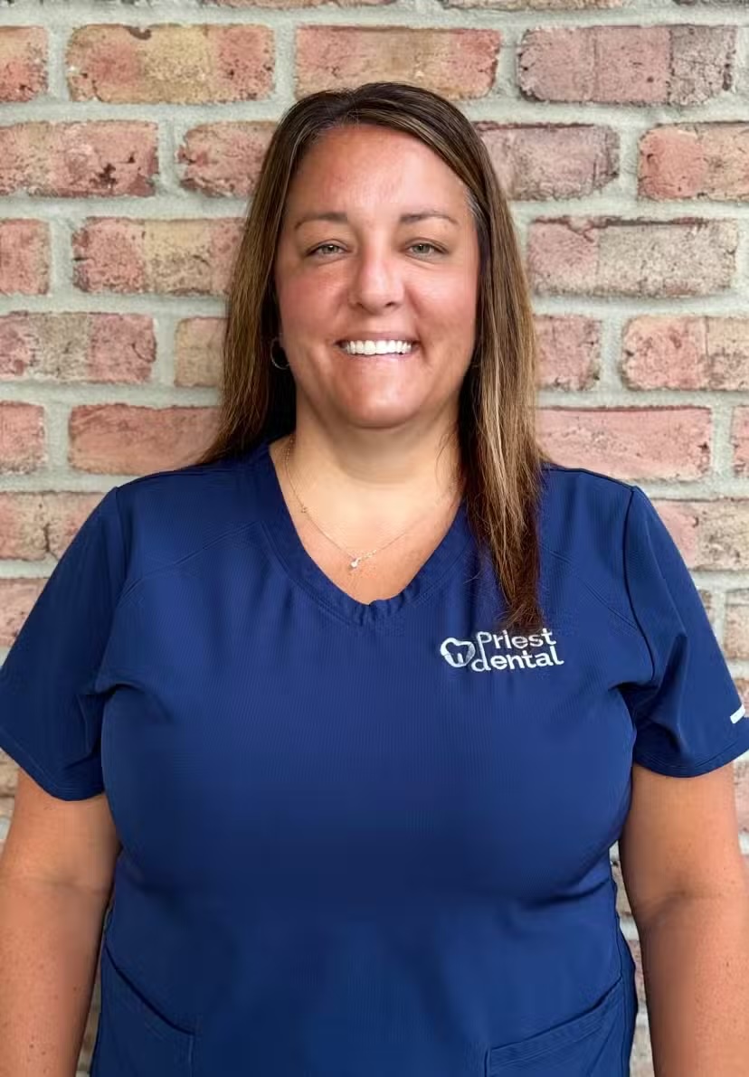 Smiling woman with long brown hair wearing a blue Priest Dental scrub top standing against a brick wall.