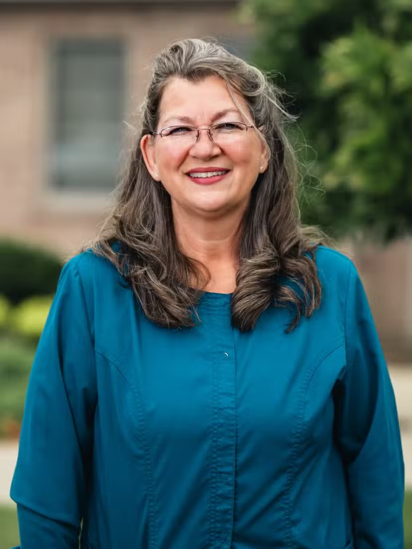 Smiling middle-aged woman with long gray hair and glasses wearing a teal blouse outdoors.