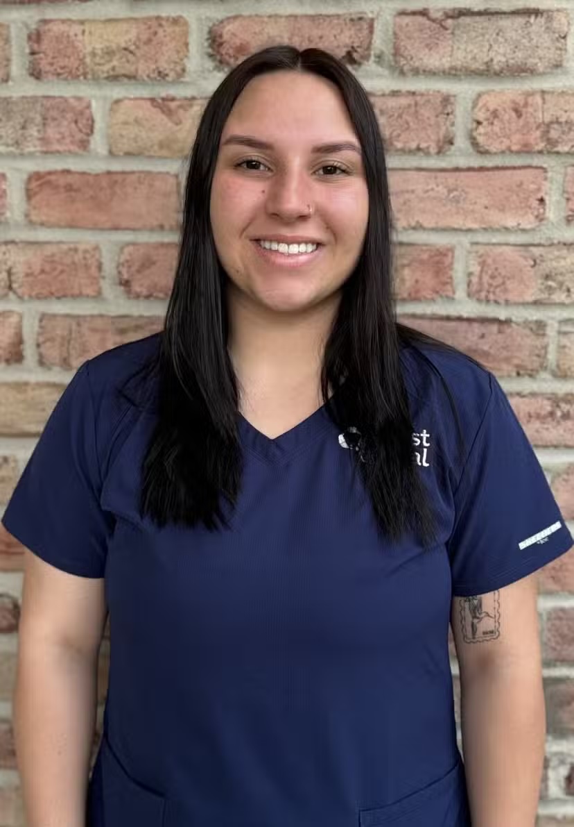 Smiling woman with long dark hair wearing a navy blue scrub shirt standing against a brick wall.