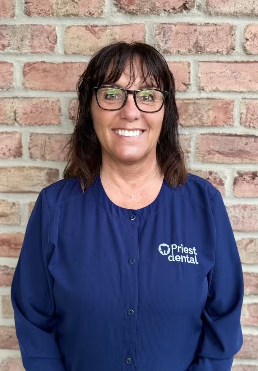 Smiling woman with glasses wearing a navy blue Priests Dental uniform standing in front of a brick wall.