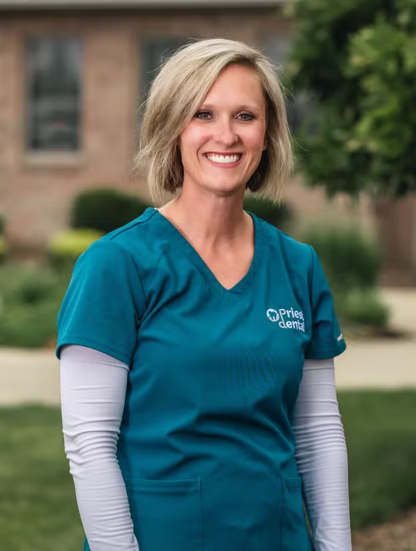 Smiling blonde woman in teal medical scrubs with white long sleeves stands outdoors in front of a blurred residential background.