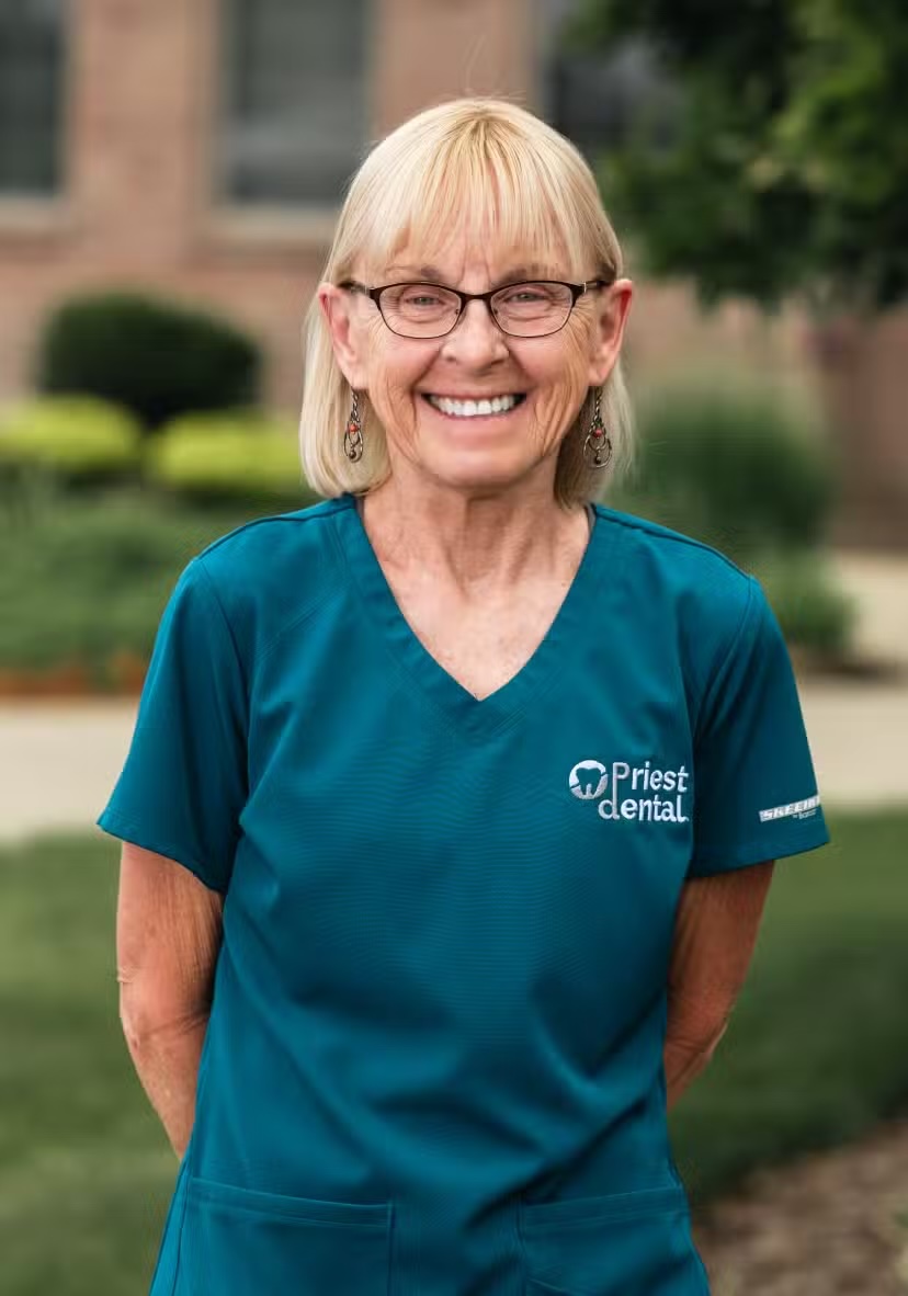 Smiling older woman wearing glasses and Priest Dental scrubs standing outdoors.