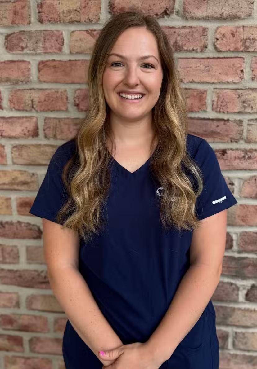Smiling woman with long wavy blond hair wearing a navy scrub top standing against a brick wall.