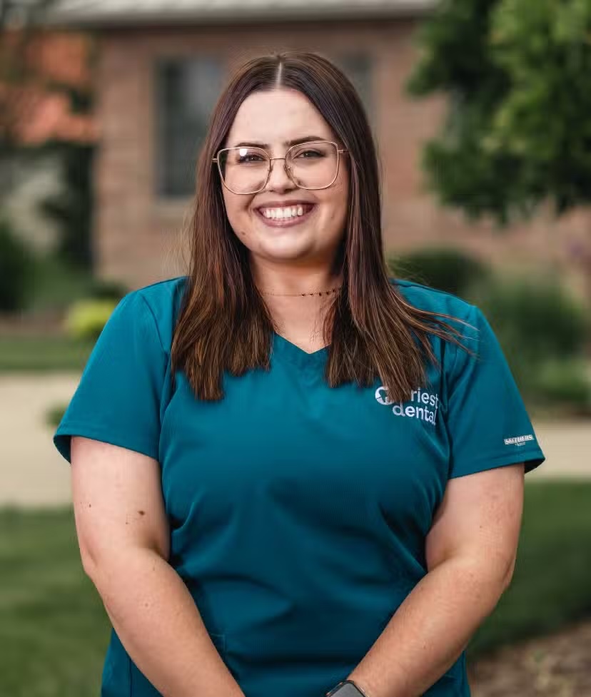 Smiling woman with long brown hair and glasses wearing a teal dental uniform standing outdoors.