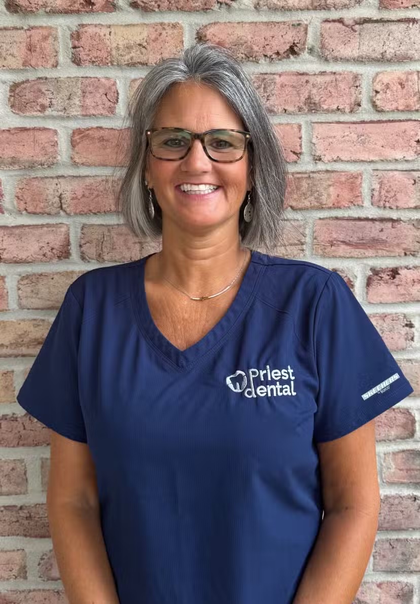 Smiling woman with gray hair and glasses wearing a navy blue Priest Dental scrub top standing against a brick wall.