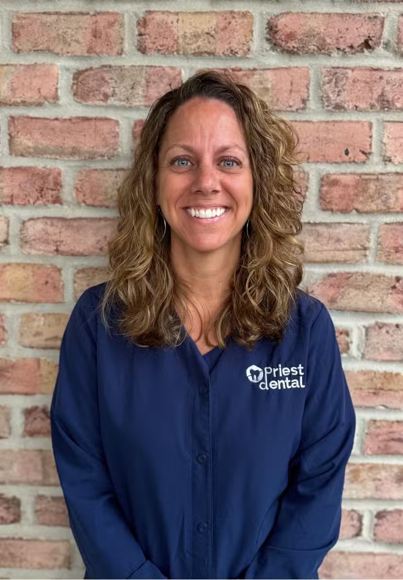 Smiling woman with curly hair wearing a navy blue shirt with the Priest Dental logo standing in front of a brick wall.