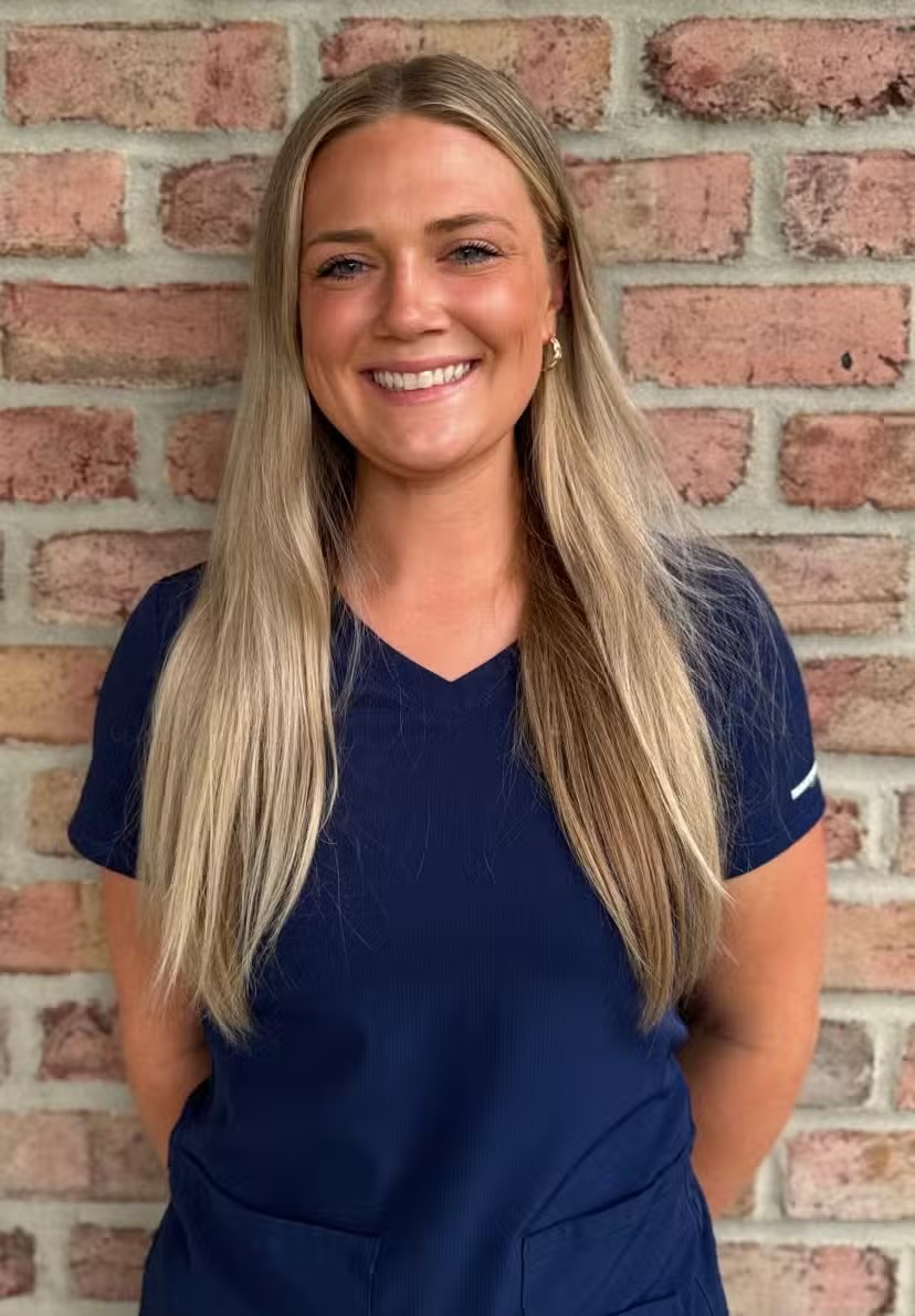 Smiling woman with long blonde hair wearing a navy blue scrub top standing in front of a brick wall.