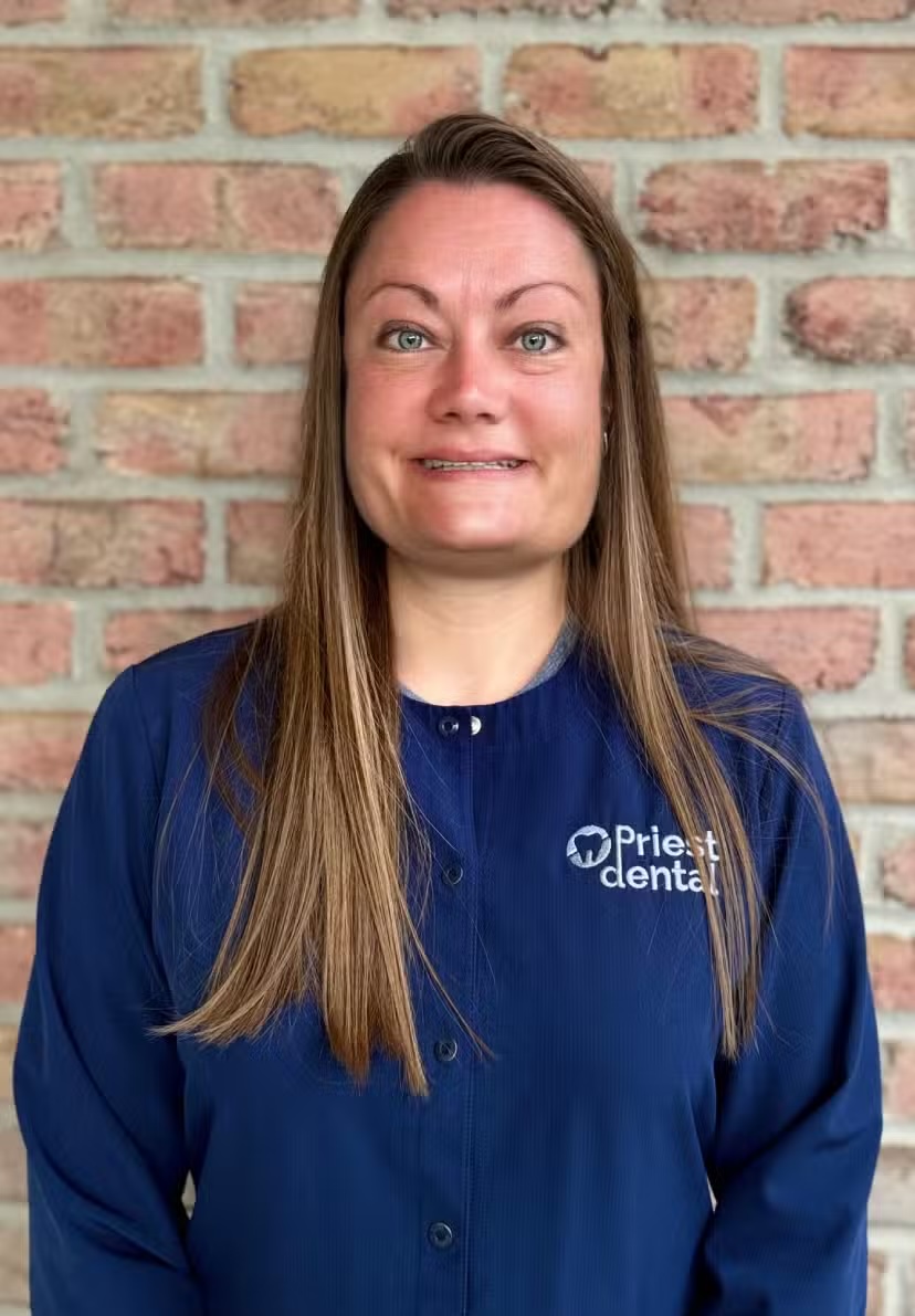 Woman with long brown hair wearing a blue Priest Dental uniform standing in front of a brick wall.