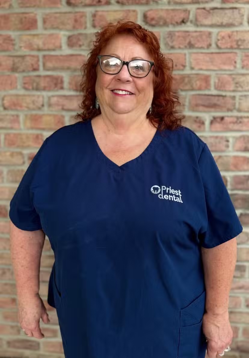 Smiling woman with red curly hair and glasses wearing a navy blue Priest Dental uniform standing in front of a brick wall.