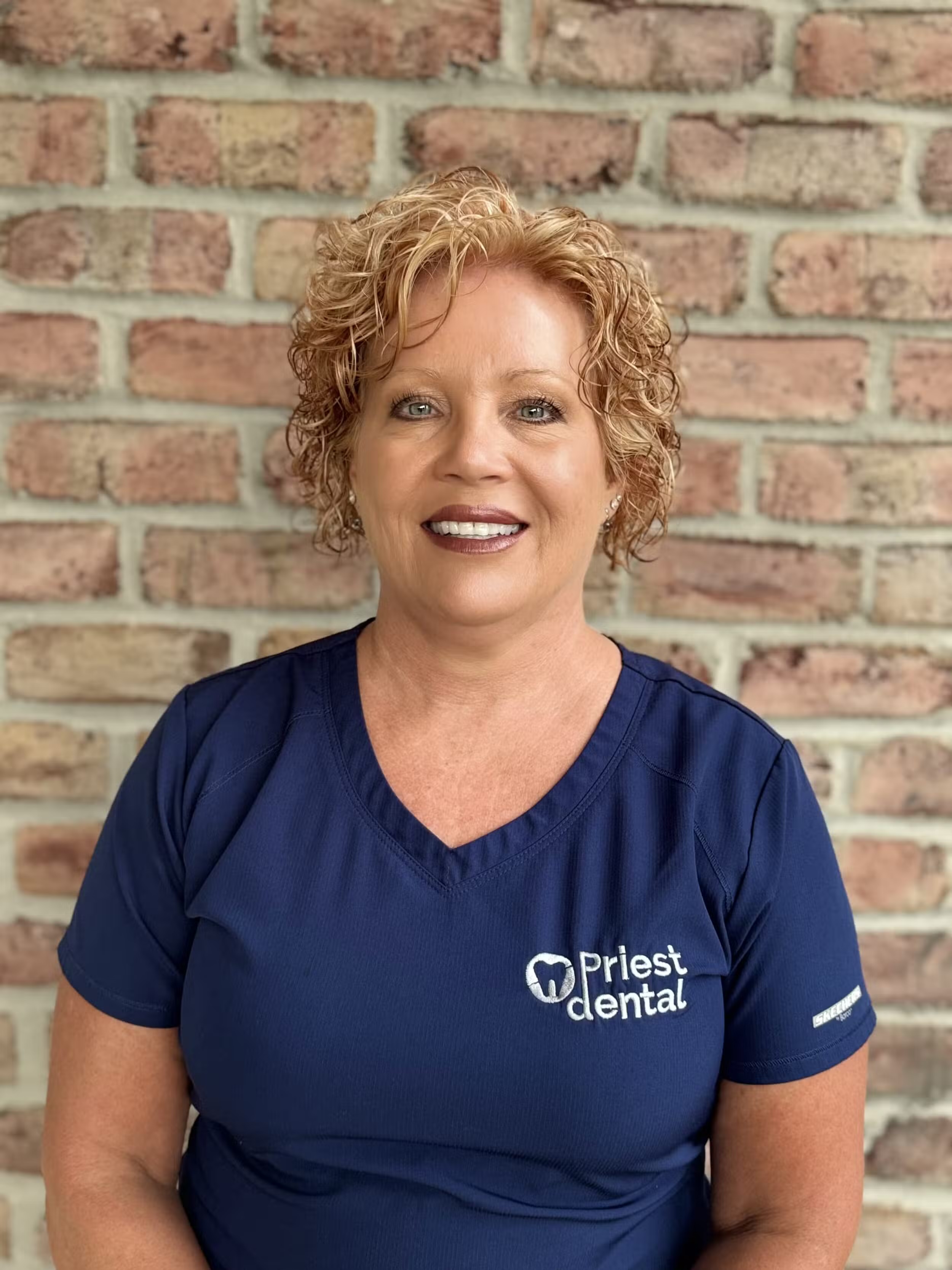 Smiling woman with short curly blonde hair wearing a navy blue Priest Dental shirt standing in front of a brick wall.