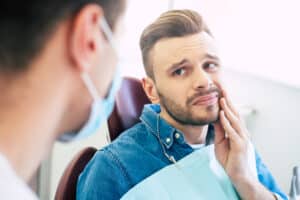 Man sitting in a dental chair holding his jaw with a pained expression, indicating possible tooth pain or dental emergency, while speaking with a dentist