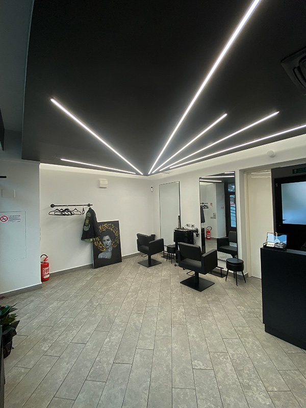 Modern hair salon interior with two black styling chairs in front of large mirrors, geometric ceiling lights, and a coat rack with a jacket and a portrait resting against the wall.