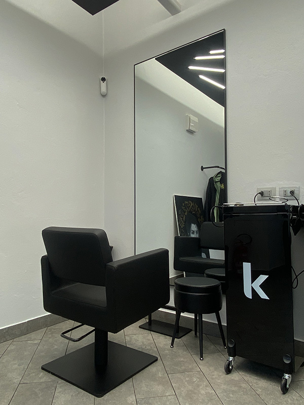 Modern salon styling station with black salon chair, large mirror, black rolling cart with letter 'k', and small black stool on gray tiled floor.