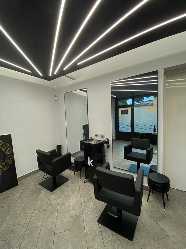 Modern salon interior with two black styling chairs, large mirrors, and sleek linear ceiling lights.