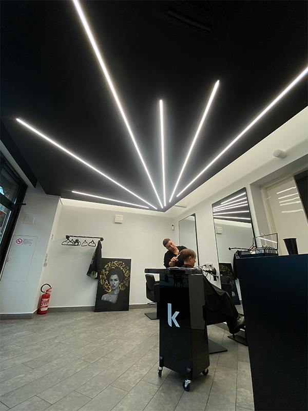 Modern barbershop interior with linear ceiling lights, a barber cutting a client's hair in front of a mirror, and a painting leaning against the wall.