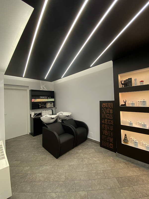 Modern salon shampoo area with two black wash chairs, white sinks, black shelves stocked with hair products, and linear ceiling lights.