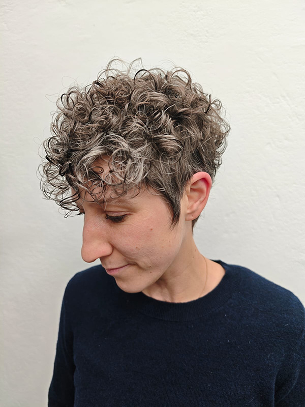 Person with short, curly brown and gray hair looking downward wearing a dark blue sweater against a plain light background.