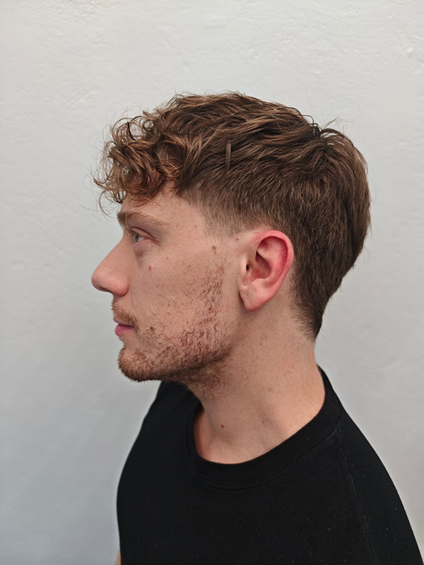 Profile of a young man with curly brown hair and light facial stubble wearing a black shirt against a plain light gray background.