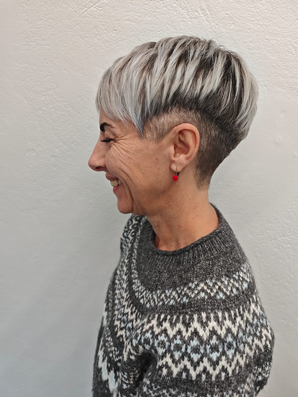 Side profile of a smiling woman with a short layered gray haircut wearing a patterned knit sweater and red earrings against a plain textured wall.