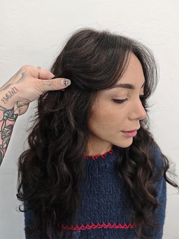 Side profile of a woman with long, dark wavy hair being styled by a tattooed hand against a plain white background.