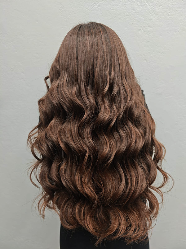 Long, wavy brown hair cascading down the back against a plain light gray wall.