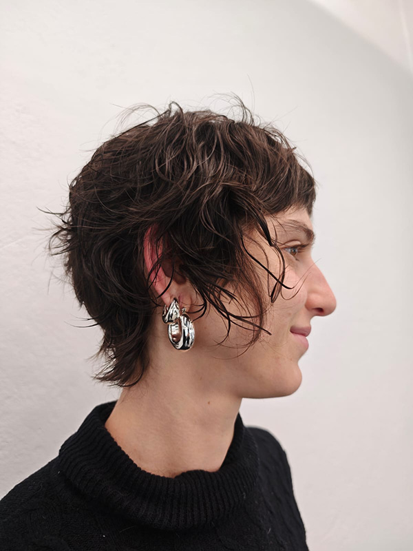 Side profile of a person with short, wavy brown hair wearing a black sweater and silver hoop earrings.